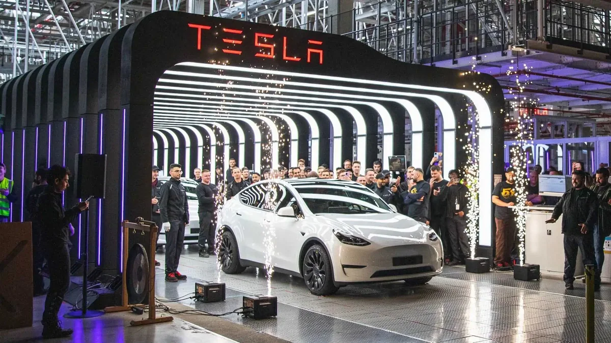 Tesla exhibition stand at an automotive show, with a white car, visitors and an illuminated Tesla sign.